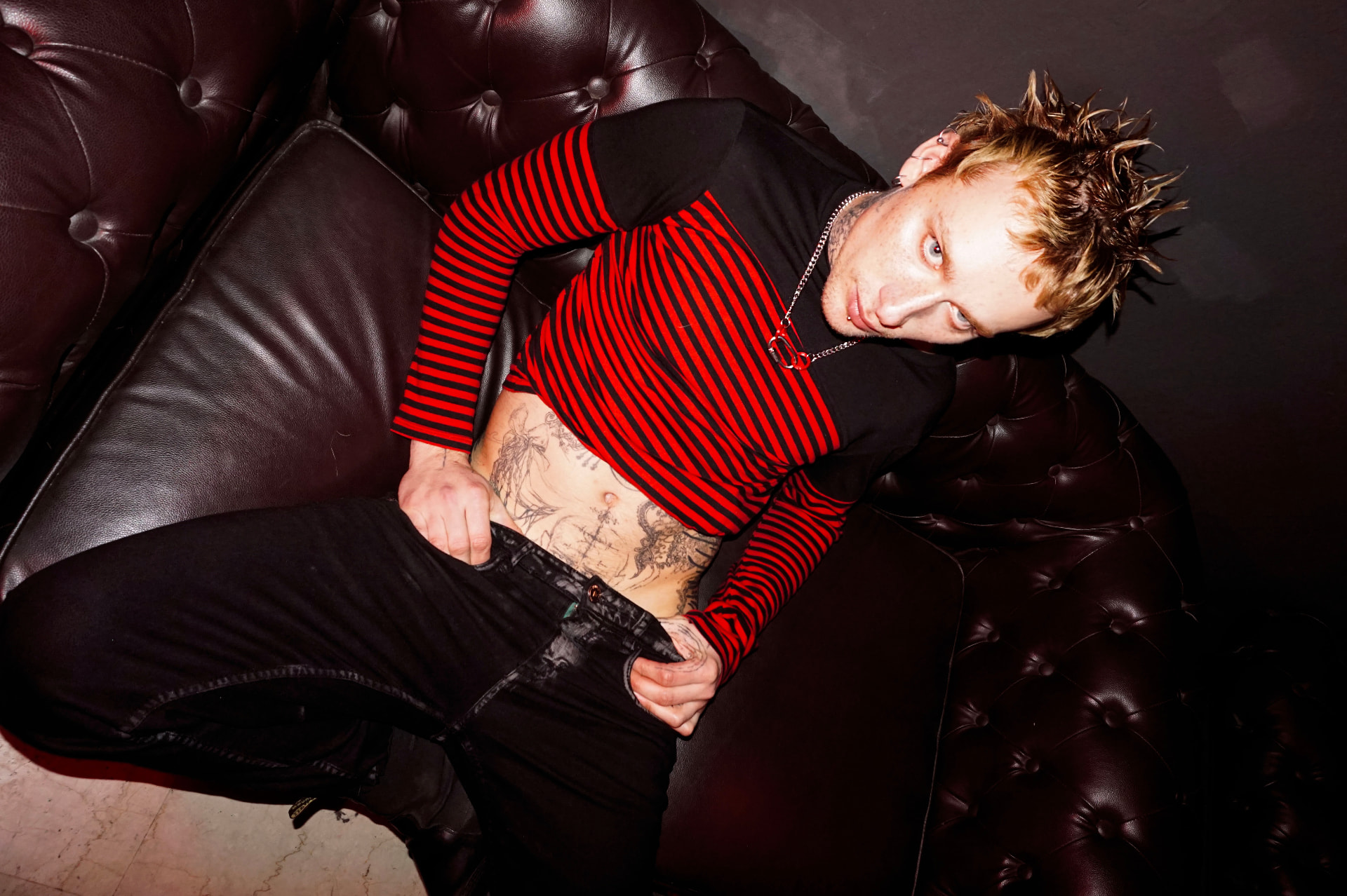 Wide editorial frame: high-angle view of the model in a red and black striped top on a dark leather sofa