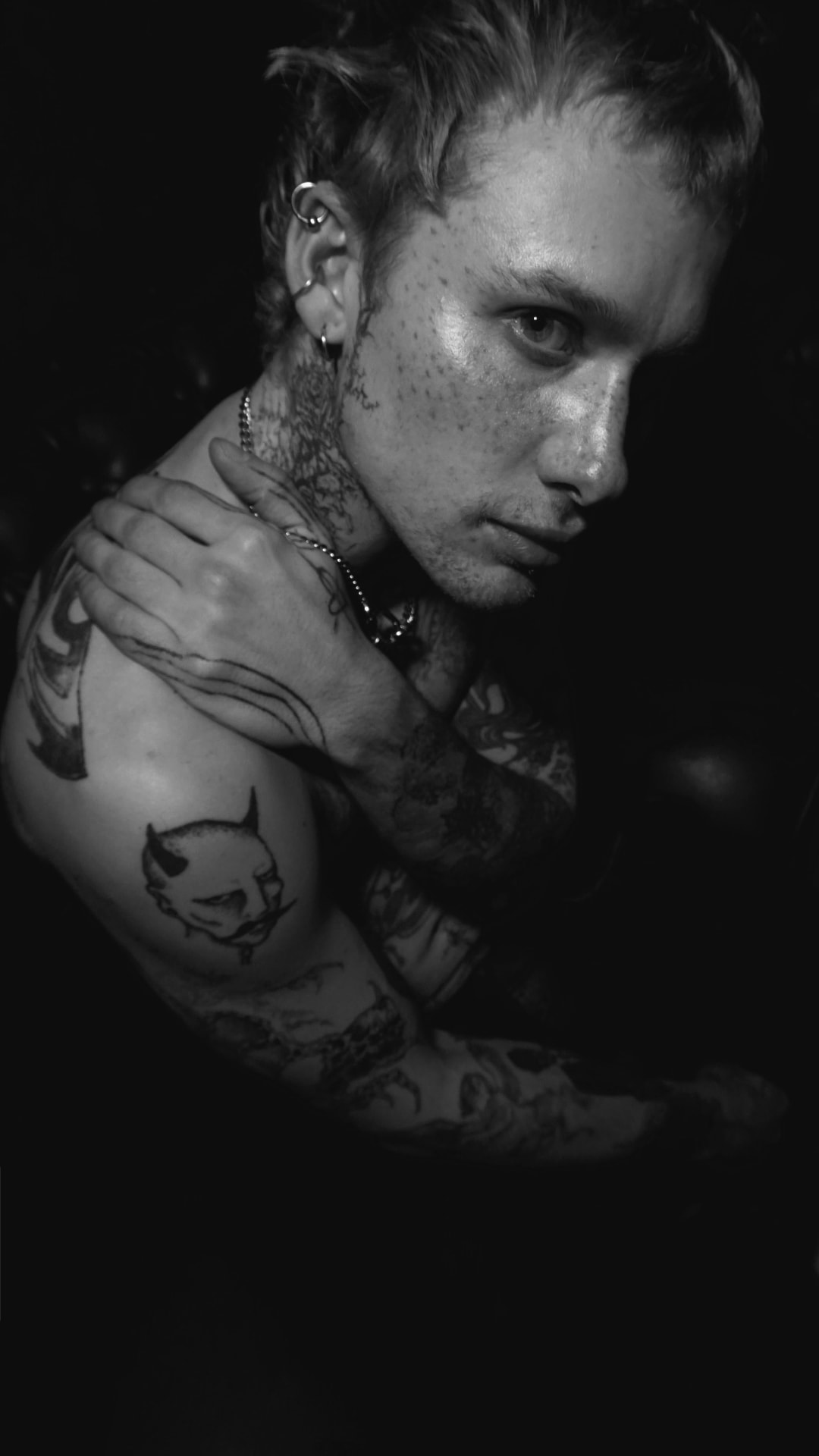 Black-and-white low-light portrait, hand resting on a tattooed shoulder, chain jewellery at the neck