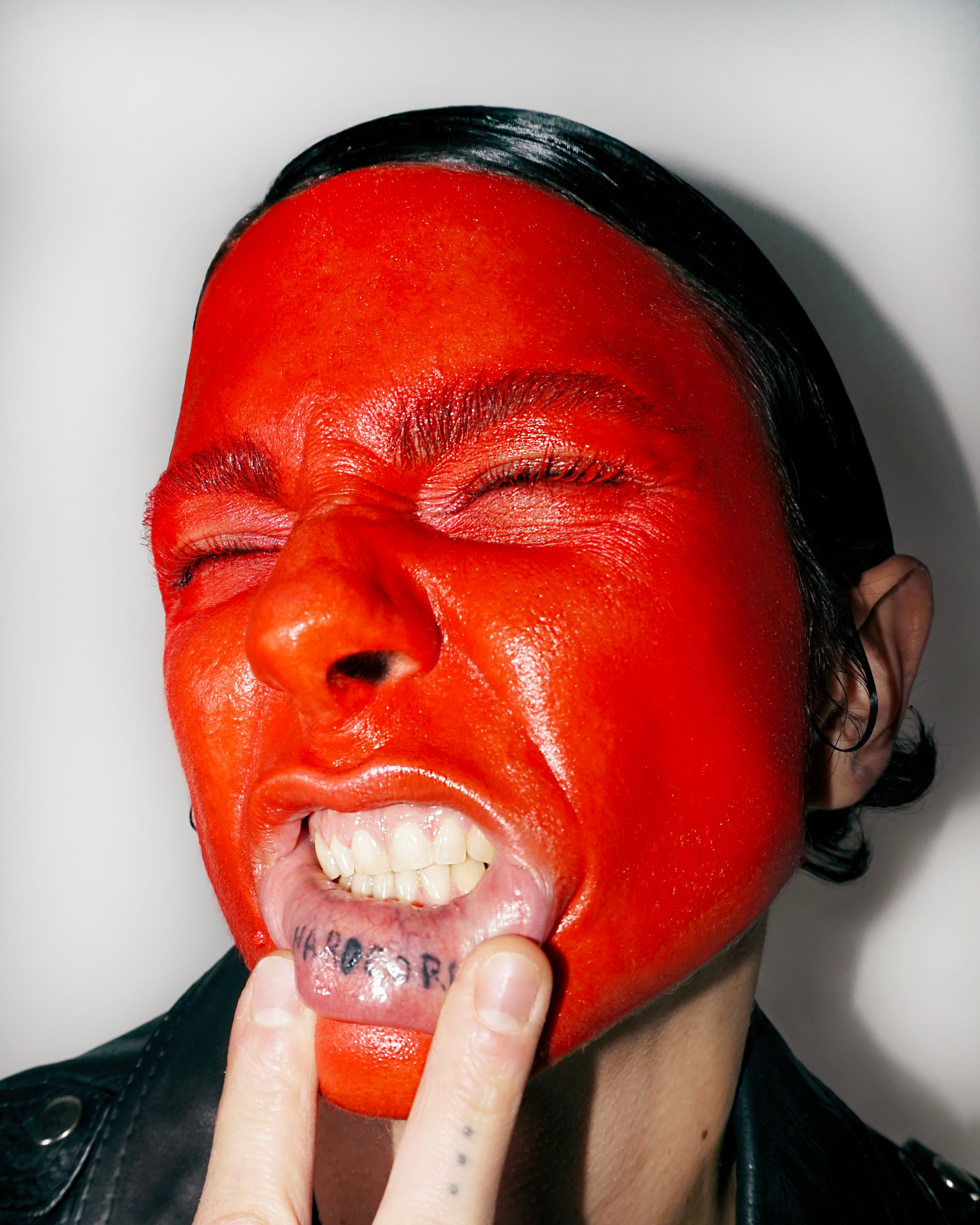 Hard-flash close-up: face fully covered in glossy signal red, teeth bared and lower lip pulled down to reveal a HARDCORE tattoo