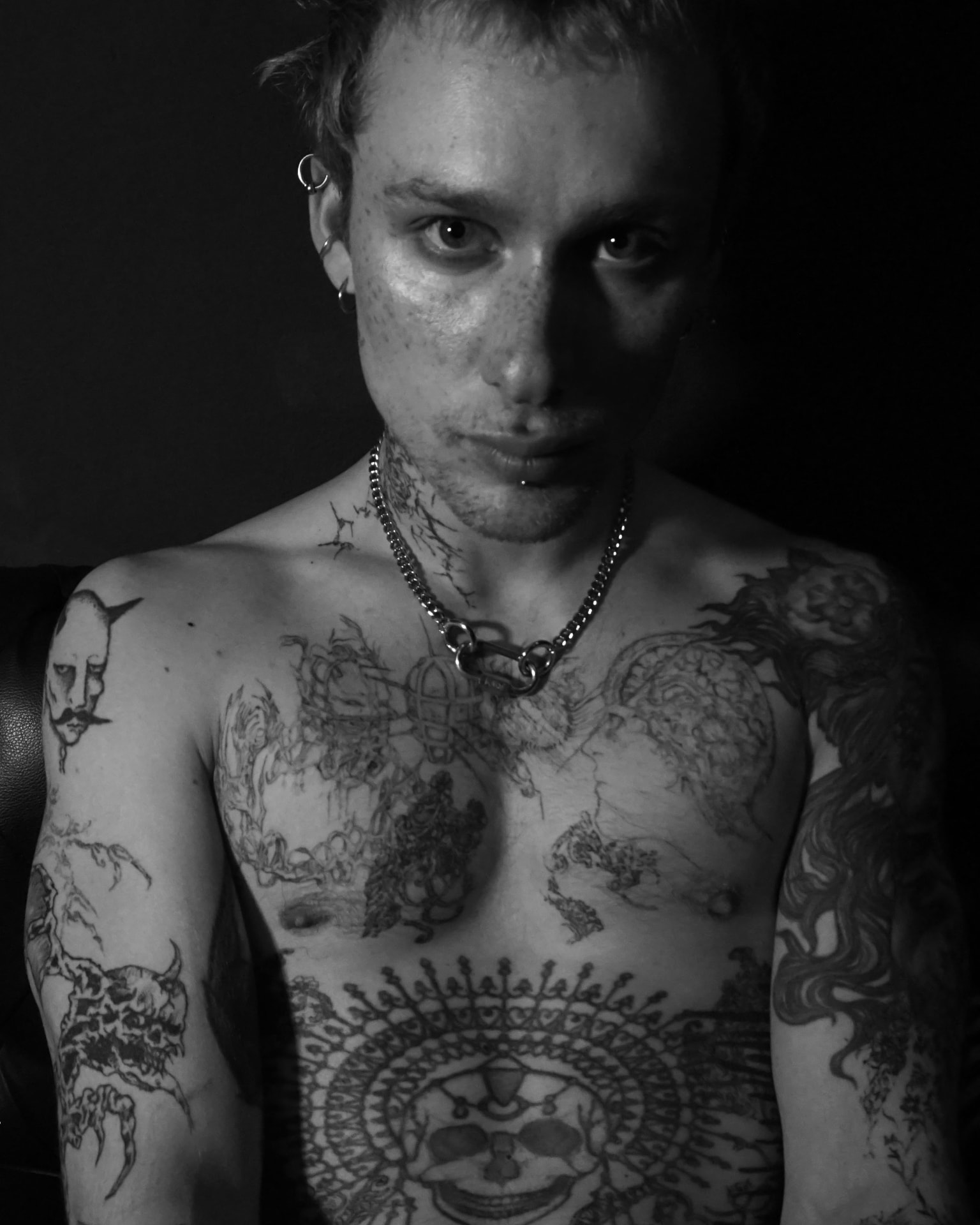 Front-facing black-and-white portrait, chain jewellery crossing the chest and tattooed shoulders