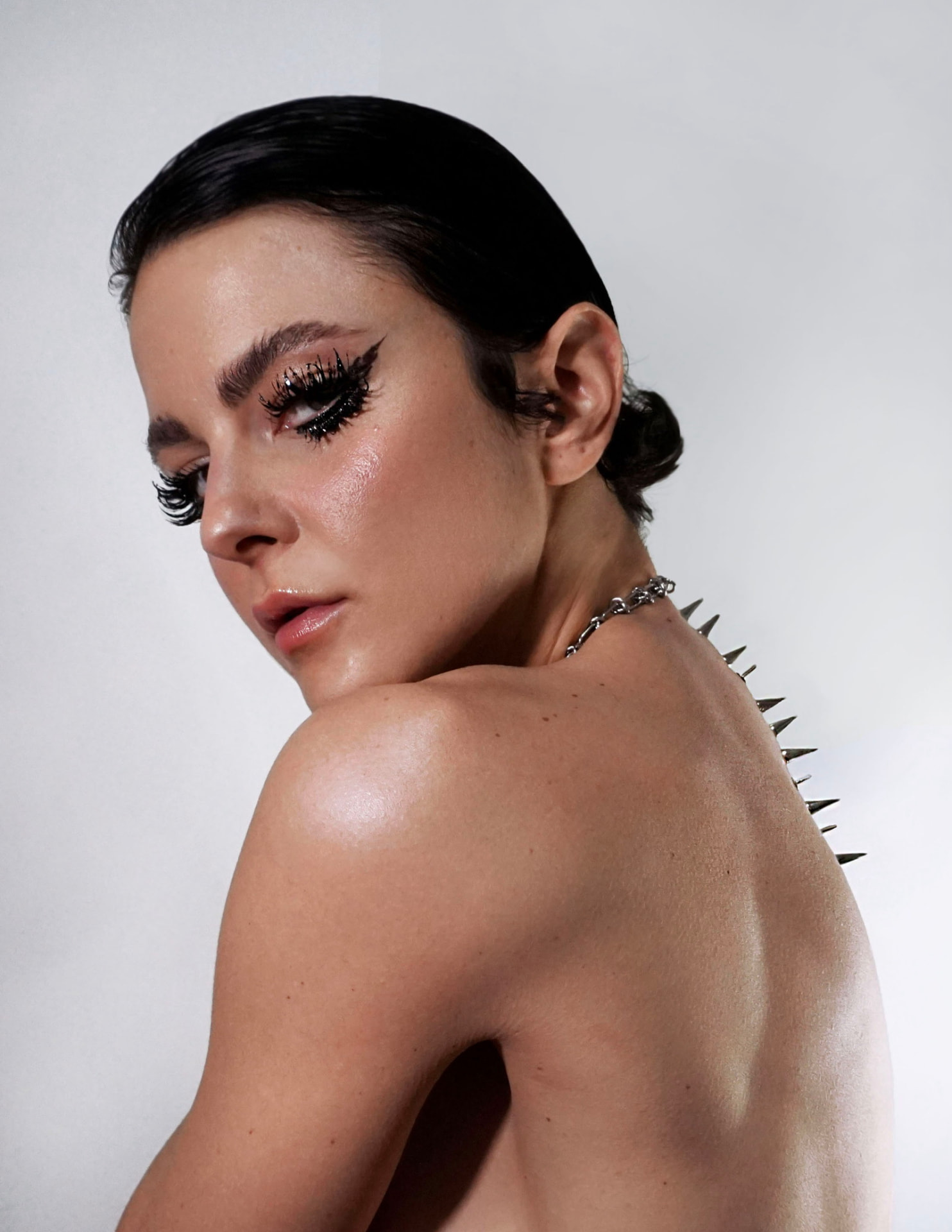 Cool-light portrait turned over the shoulder: graphic black liner, glossy skin, chain necklace and a row of metal spikes along the spine