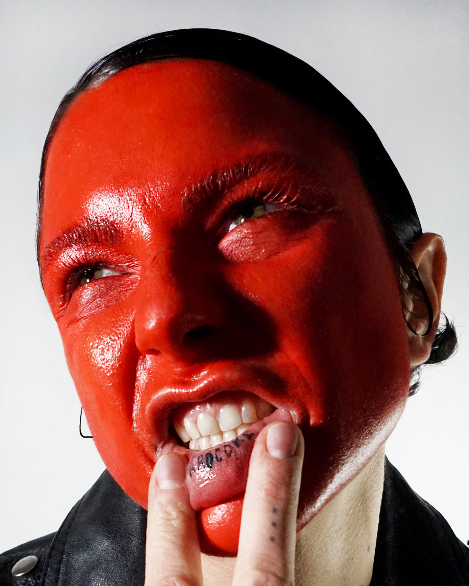 Hard-flash portrait: face covered in glossy signal red, finger pulling the lower lip down to reveal a HARDCORE tattoo